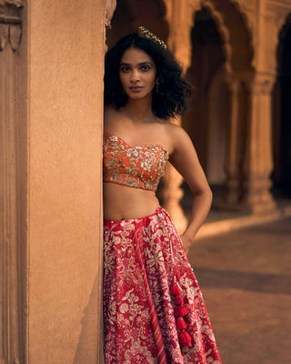 A pink multi-tone lehenga, featuring a delightful combination of leheriya and ornamental signature floral prints that evoke the charm of the ancient craftsmanship of Rajasthan. It is paired with an intricate hand–embroidered corset blouse in sunset orange, and a delicate hand–embroidered scallop dupatta.